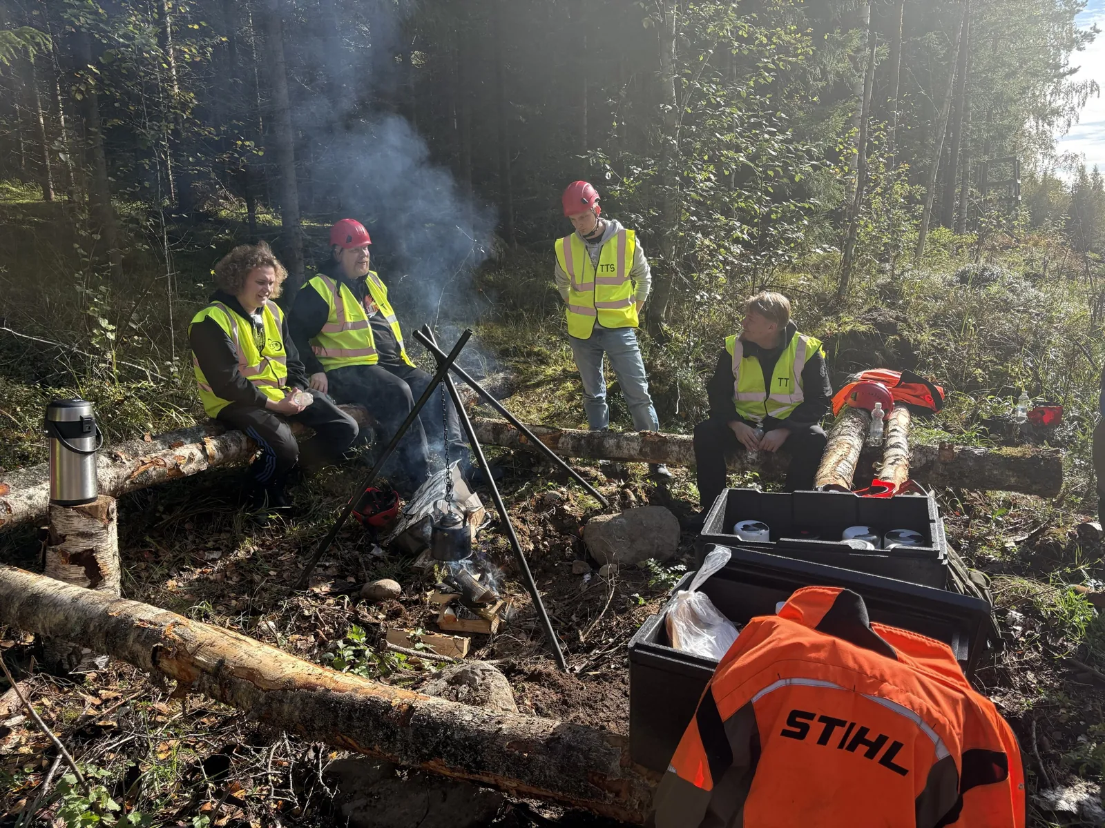 Nokipannu kolmijalalla nuotion yllä, porukka istuu kaatuneilla rungoilla.